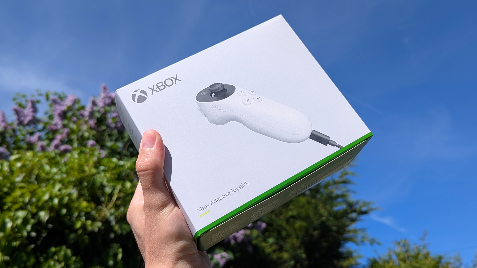 Адаптивный джойстик Xbox в розничной коробке, в руке на фоне голубого облачного неба