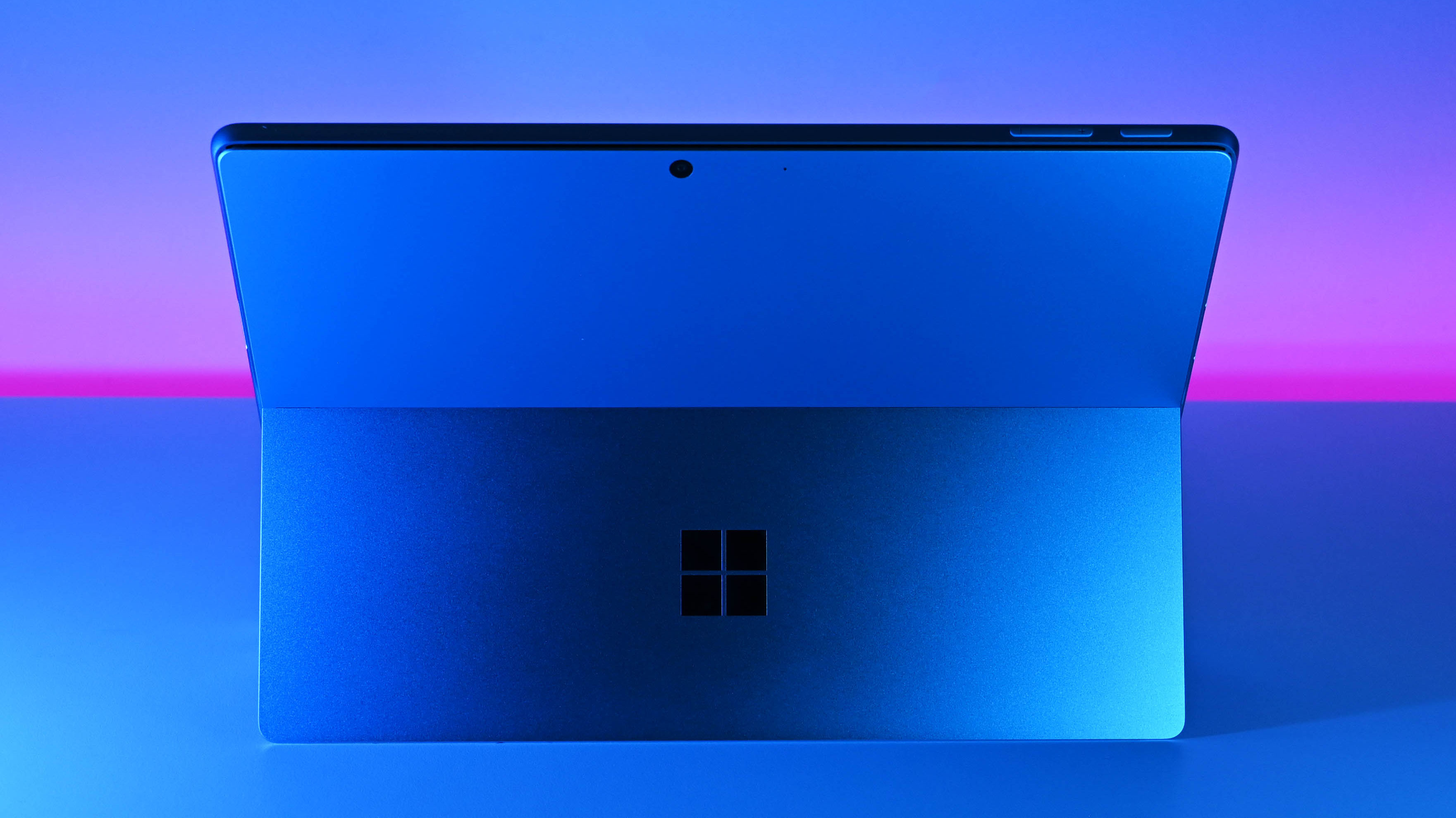A Microsoft Surface Pro 11 tablet is shown from the back in dramatic blue and pink lighting, highlighting its sleek design and iconic logo, against a gradient background.