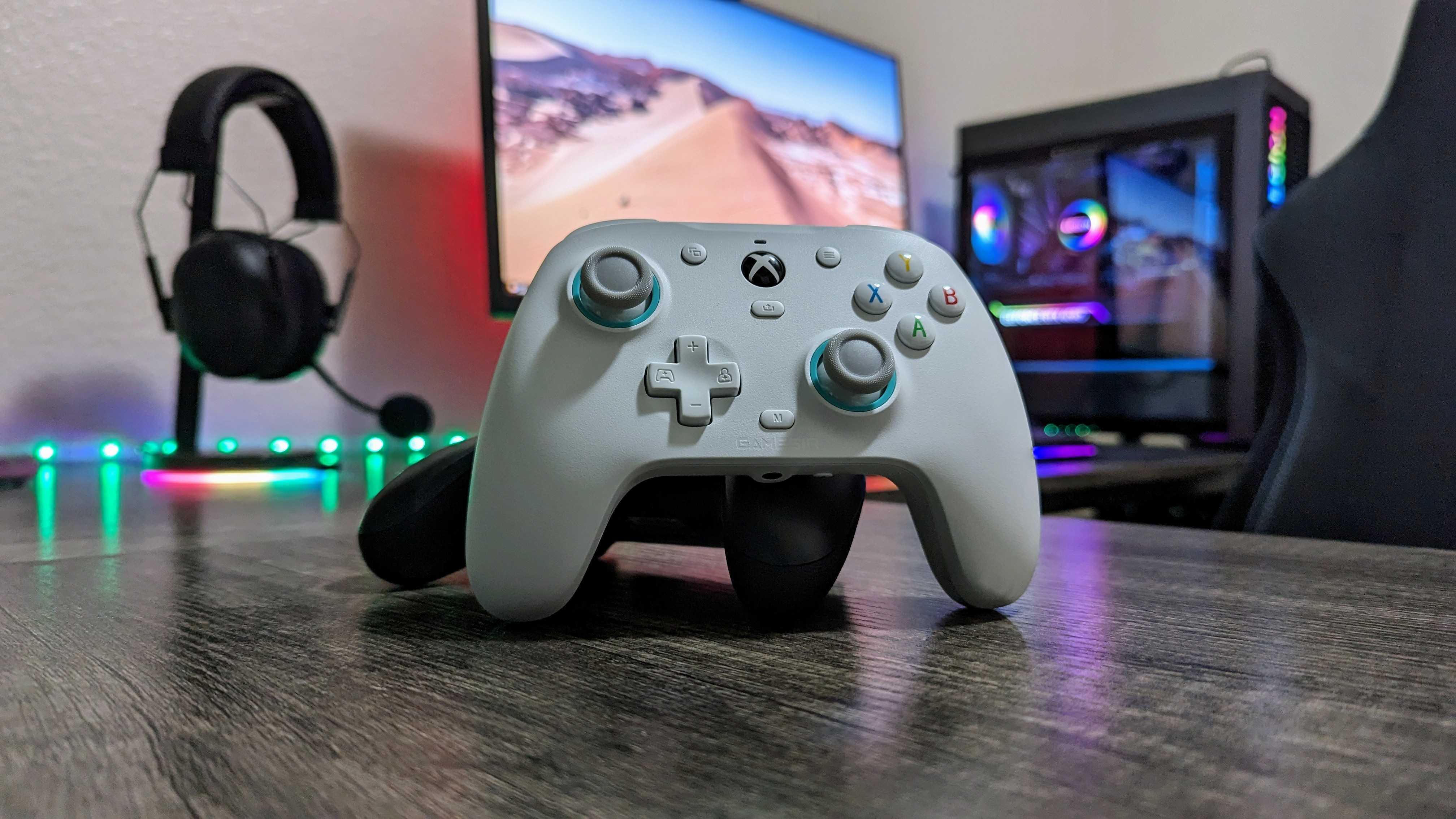 Gaming setup with a white Xbox controller on a wooden desk, against a backdrop with a monitor displaying a desert scene, headphones hung on the wall, and a glowing gaming PC.