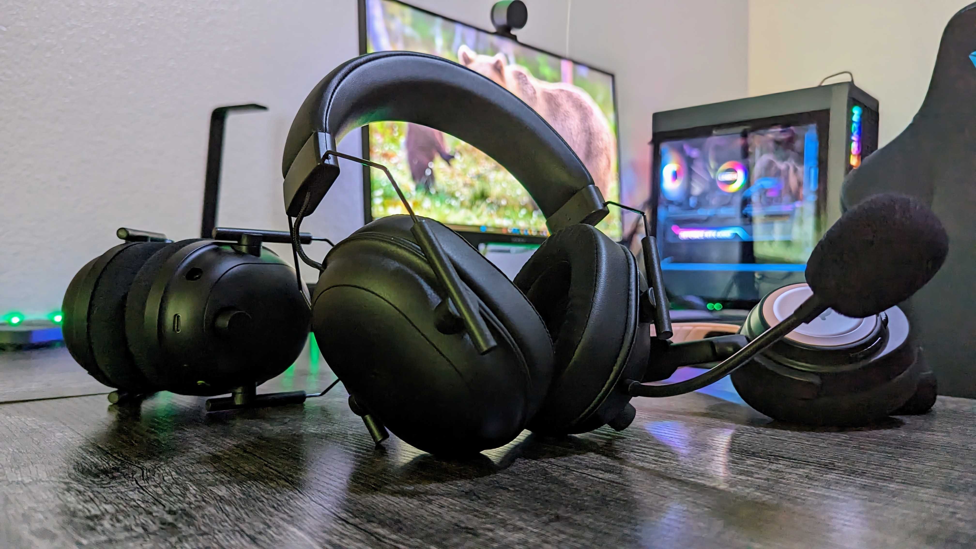 A pair of black gaming headsets with attached microphones rest prominently on a desk, surrounded by a curved monitor displaying nature and a lit gaming PC.