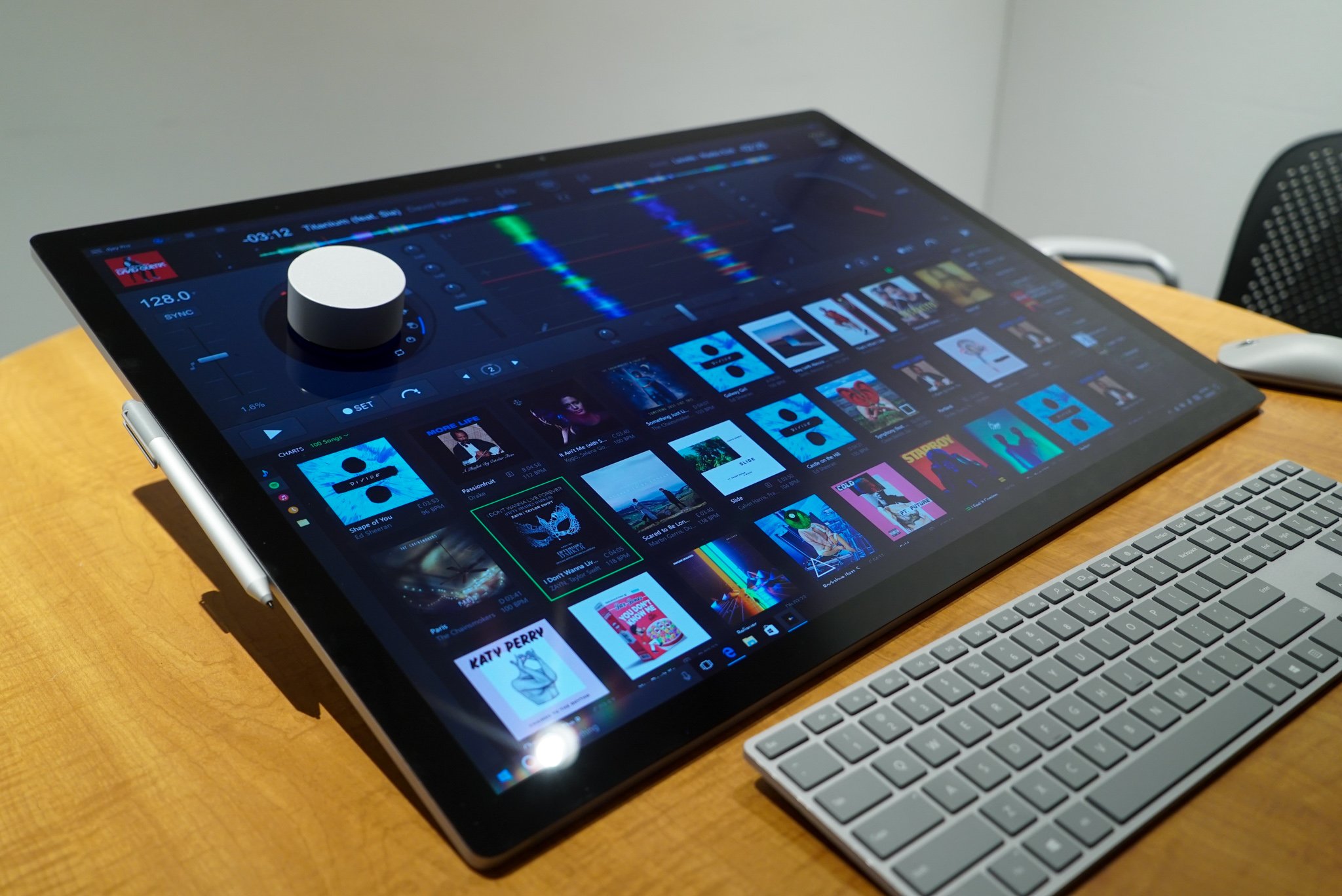A large touchscreen monitor on a wooden desk displays a music library interface. Nearby are a white stylus, a dial, and a compact keyboard.