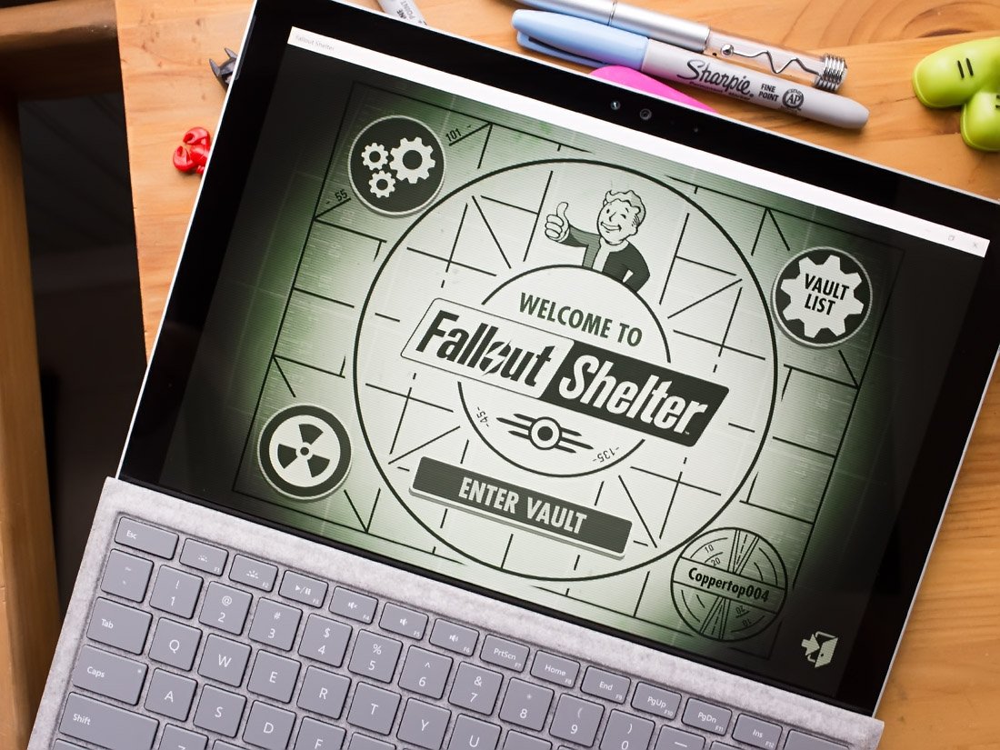 Laptop displaying the "Fallout Shelter" game main screen with a cartoon character giving a thumbs-up. Nearby are markers and game controllers.