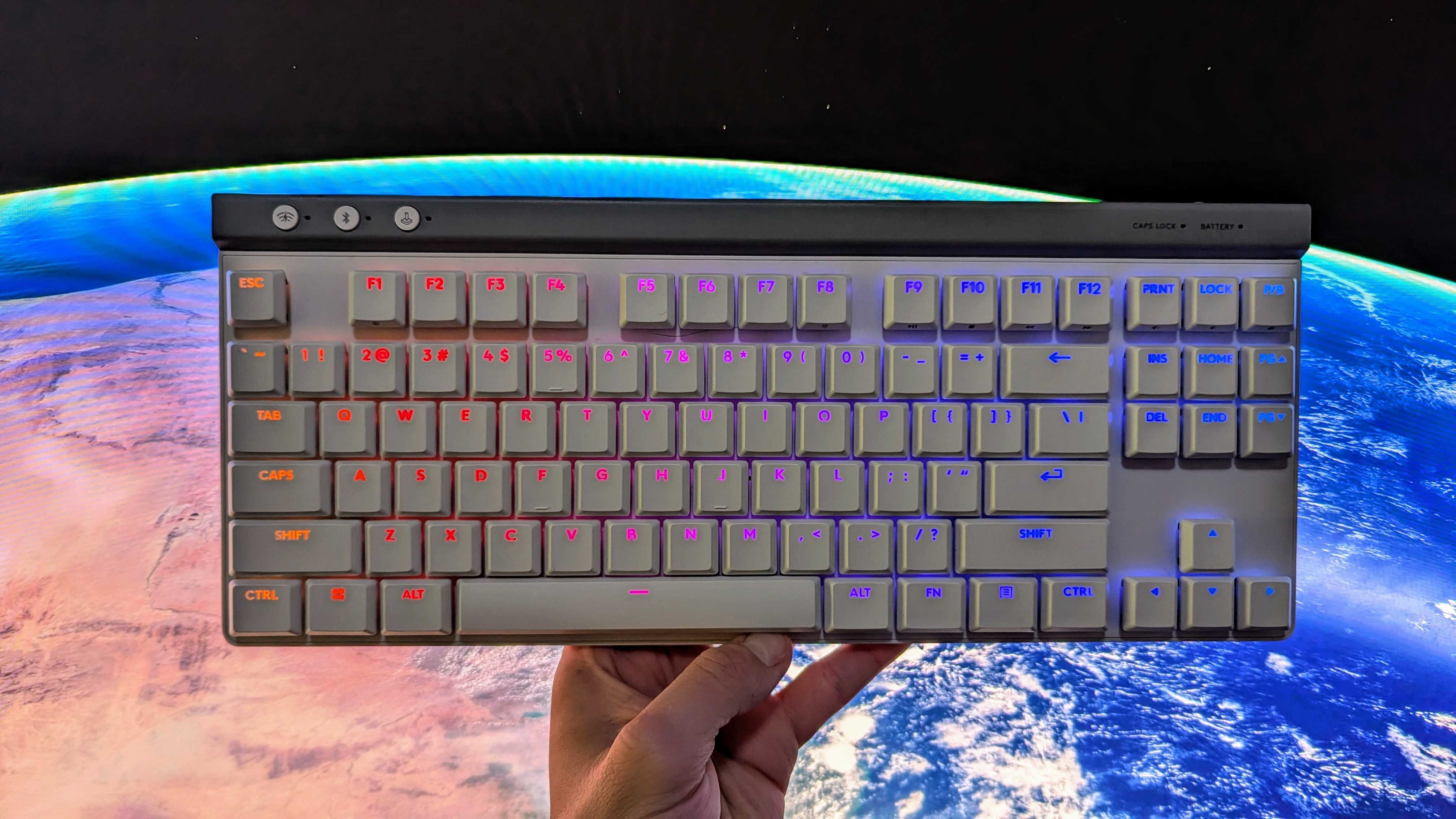 A hand holds a white mechanical keyboard with multicolored backlit keys against a dramatic Earth-from-space background, creating a futuristic feel.