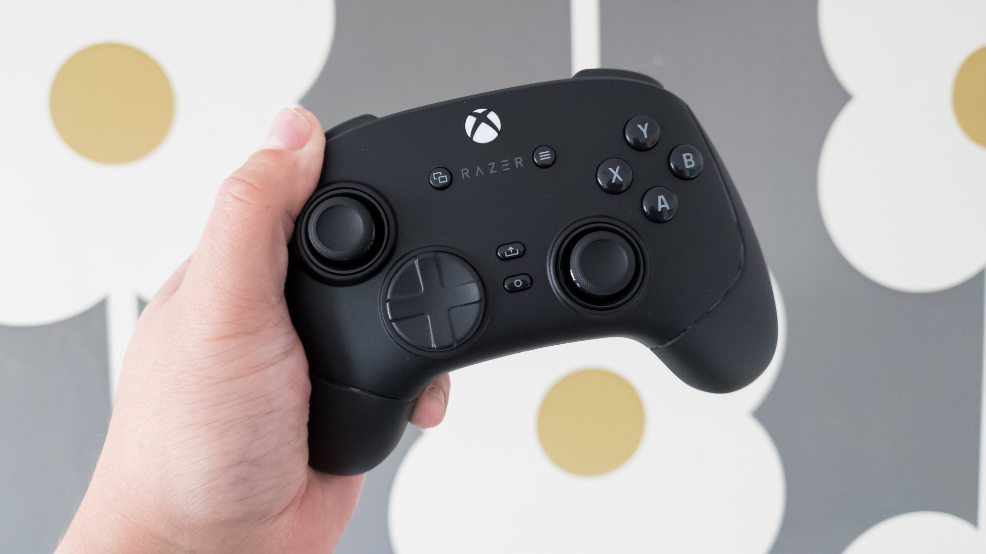 A hand holds a black Razer gaming controller against a background with large, abstract flowers in gray and white with yellow centers.