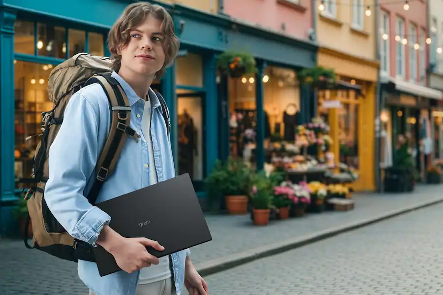 Young person with a backpack holds a laptop, walking down a charming cobblestone street lined with colorful shops and flower displays, conveying exploration.