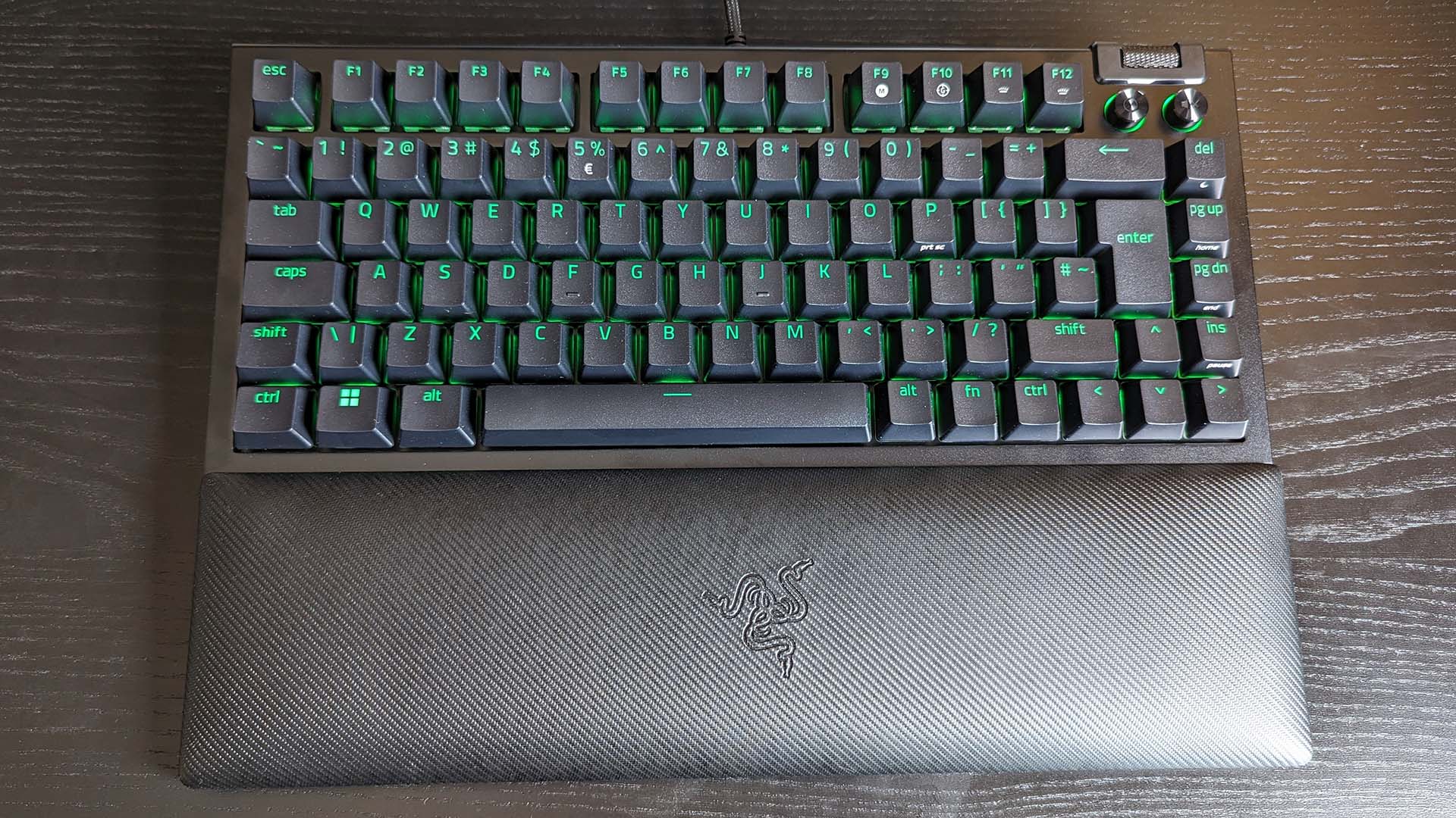 Mechanical keyboard with green backlighting on black keys, placed on a dark wood surface. Includes a textured wrist rest with a logo.