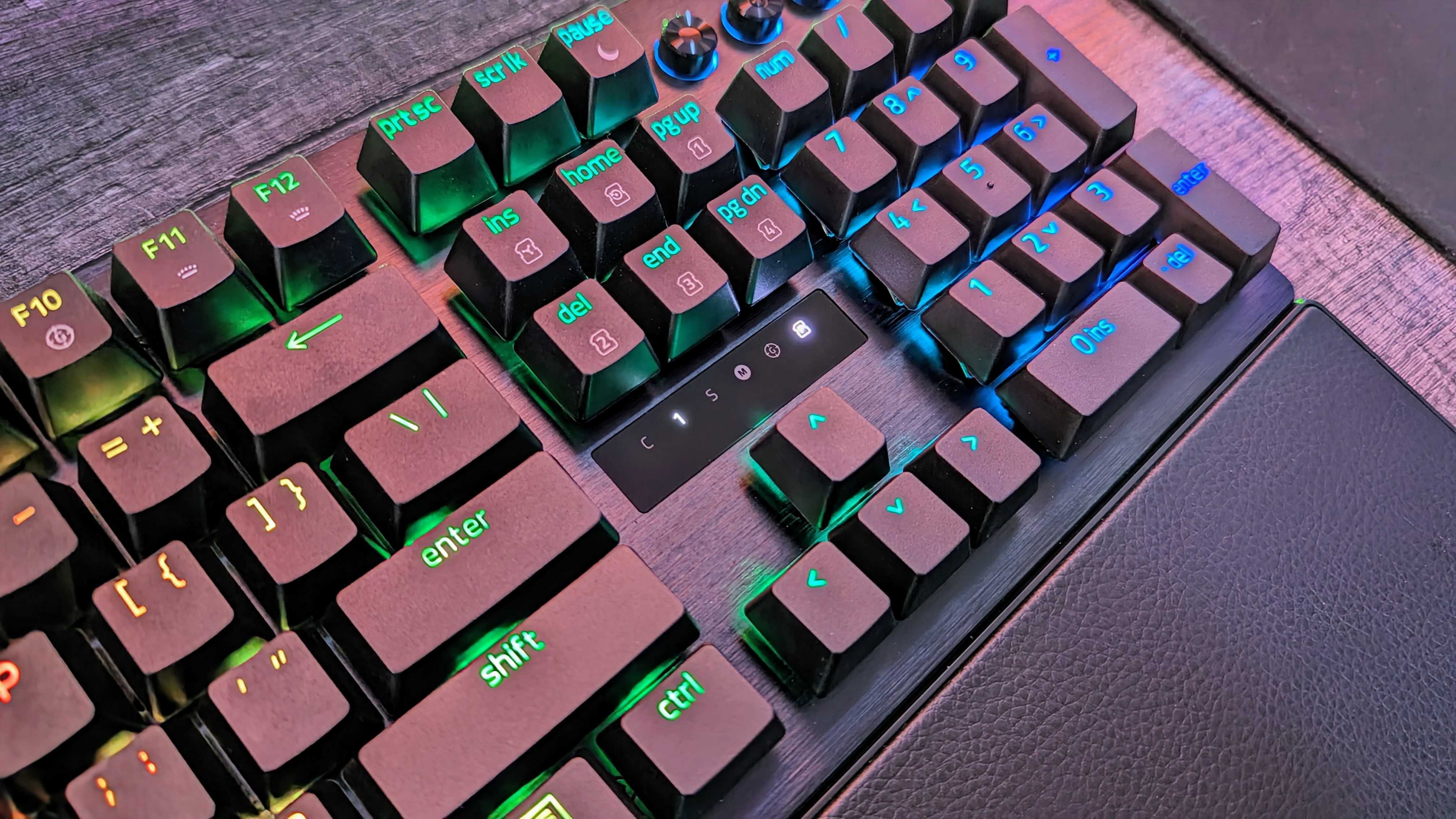 Mechanical keyboard with RGB lighting on wood surface. Keys are illuminated with green and blue hues. Includes a wrist rest. Tech and gaming vibe.