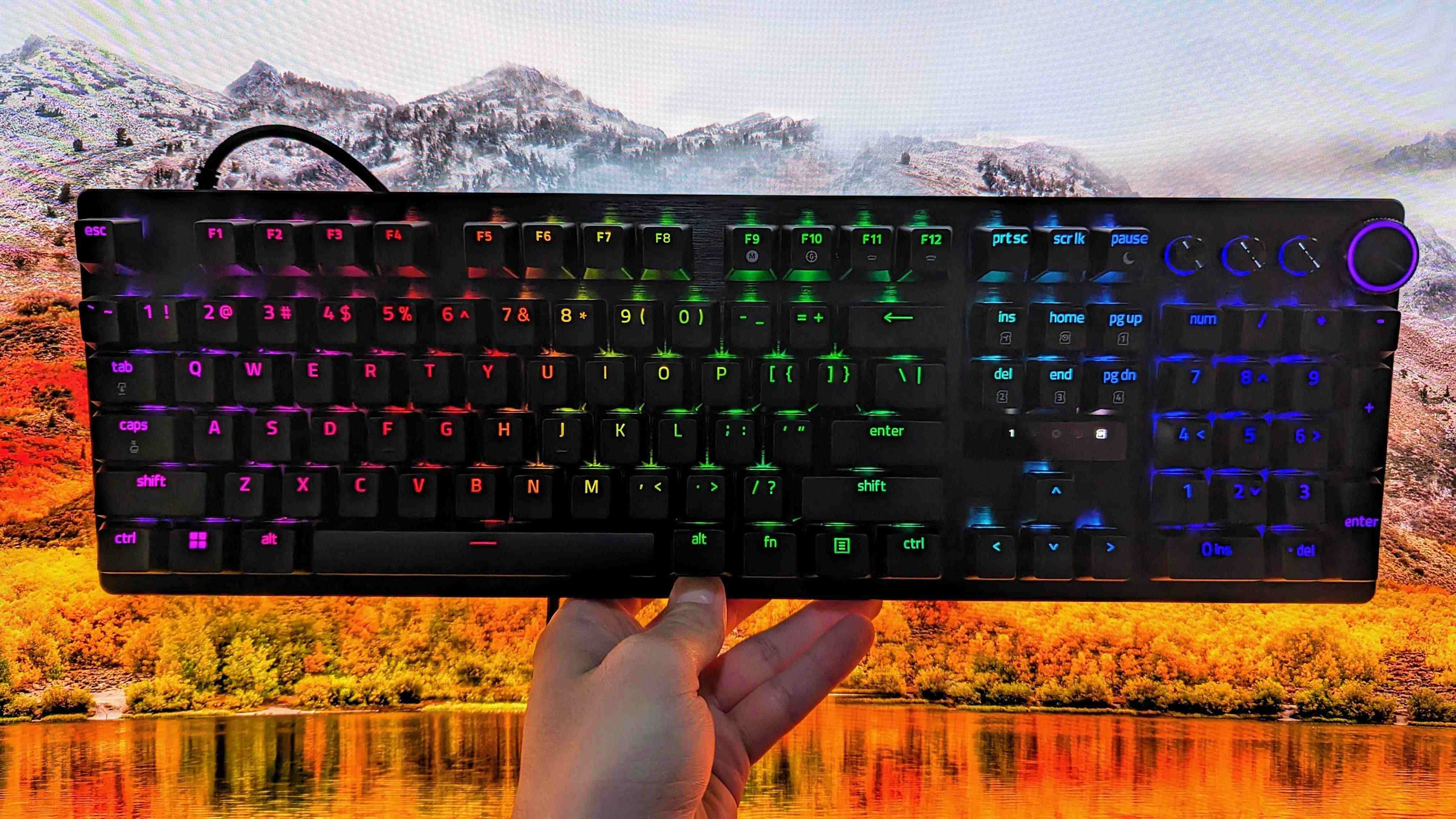 A hand holds a black mechanical keyboard with multicolor backlit keys in front of a vibrant autumn landscape, featuring mountains and a lake.