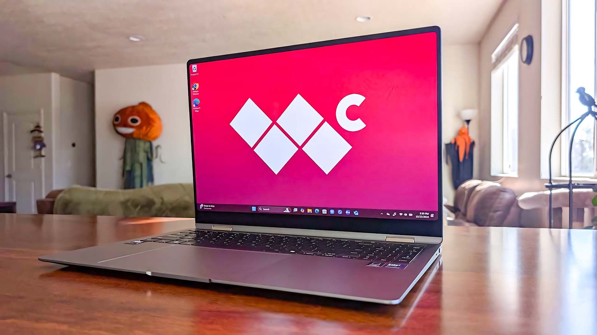 A sleek laptop open on a wooden table shows a red screen with a minimalist logo. In a cozy living room, a quirky, child-sized pumpkin figure is in the background.