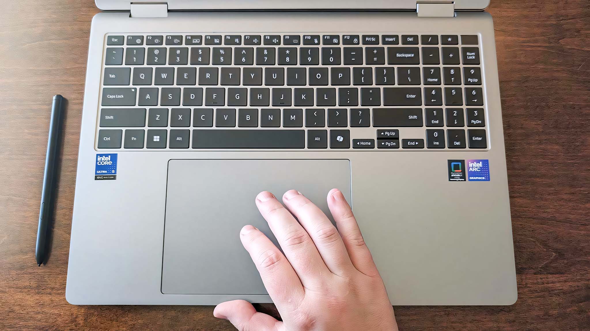A hand rests on the touchpad of a silver laptop with a black keyboard. A stylus is placed on the wooden surface beside it. The scene conveys focus.
